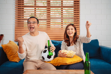 Asian Couple watching sport football cheering excited on sofa in living room at home. enjoy relaxing happiness.