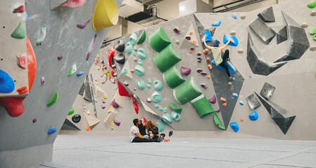 Multi-Cultural Group Watching Friend On Climbing Wall In Indoor Activity Centre