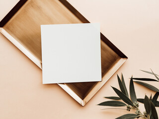 White blank paper square card mockup on gold tray and green eucalyptus branch on beige background.