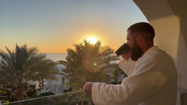 Dreaming european middle aged bearded man in bath robe stands in the balcony with great views to the hotel, palms, sea, sun, and drinks hot coffee or tea, sun shine, enjoying beautiful morning