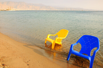 Fototapeta na wymiar Eilat, Israel. Beach. Two plastic empty chairs and view of wooden ship in the sea and mountains at background. Winter sun escape destination.