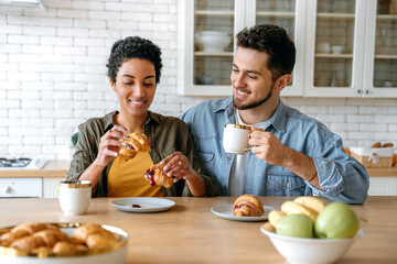Fototapeta na wymiar Morning breakfast. Happy multiracial couple, caucasian man and african american woman, spend time together at home, they drink coffee, eat croissants, chat on different topics, plan leisure time,smile