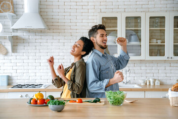 Joyful happy family couple, african american woman and caucasian man, having fun and dancing together at home in the kitchen while preparing lunch, smiling. Happy leisure time together