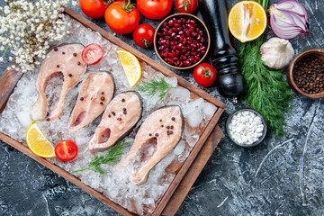 Fototapeta na wymiar top view raw fish slices with ice on wood board bowls with pomegranate seeds sea salt black pepper onion garlic dill tomatoes on grey background
