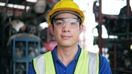 Portrait of young Asian man warehouse worker wearing safety glassed and safety uniform looking to camera with arms crossed while standing at old motor automotive spare parts warehouse