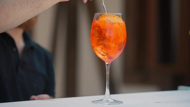 Barman is pouring champagne into aperol spritz cocktail glass with orange slices. Some seashells, towel and sunglasses near swimming pool. Vacation, summer, holiday, luxury resort concept