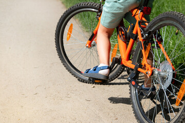 photo of a child riding an orange bicycle in a city park. A warm summer day. Bottom and back view....
