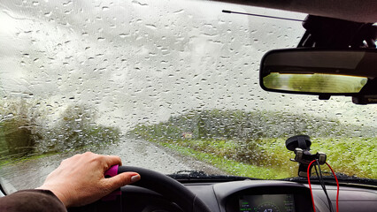 View from the car windshield from the driver's seat and hand on the steering wheel of woman on during rain, drops on the glass and nature. Travel in rainy autumn, spring or summer weather - obrazy, fototapety, plakaty