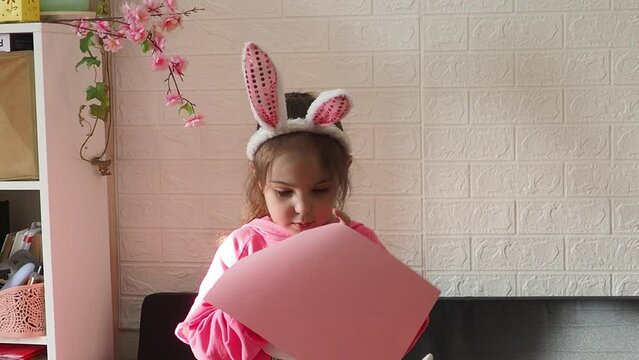 a little cute girl in a pink dress cuts out hares from paper at home. preparing for Easter. daughter painting eggs. Happy family preparing for Easter. Cute little child girl wearing bunny ears