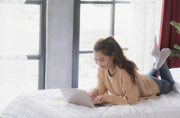 Asian woman in brown sweater lay down on bed, typing, chatting, using laptop with smile of fun and happiness, online working, networking connection concept.