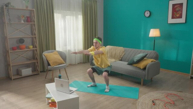 Young man doing situps on a gym mat in the living room. A man watches a video lesson on a laptop and repeats the exercises. Home fitness concept. Slow motion. HDR BT2020 HLG Material.