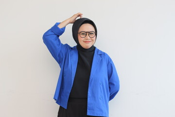 Beautiful young Asian Muslim woman, wearing glasses and blue blazer is scratching head with confused expression while smiling