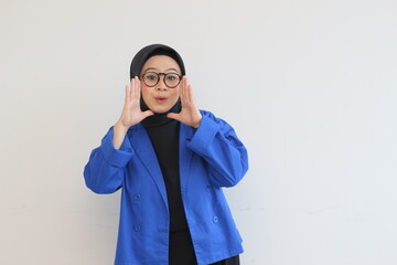 beautiful young Asian Muslim woman, wearing glasses and blue blazer in whispering gesture while smiling