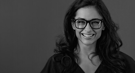 Beautiful thinking toothy smiling brunette business woman looking happy in eye glasses with curly hair style dark grey background with empty copy space. Closeup portrait. Black and white