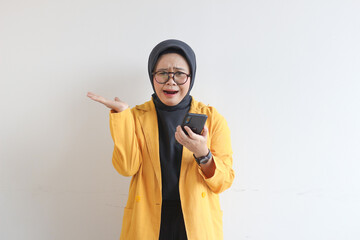 beautiful young asian woman in glasses, hijab and wearing yellow blazer looking at her mobile phone...
