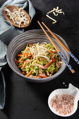 Side view vertical shot of a delightful chicken dish in an Asian style bowl. It's made with vegetables, Peking cabbage, soy sprouts, and a touch of pink sea salt, served on a black background.