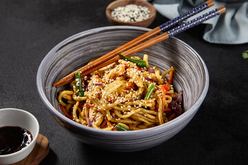 Fresh wheat noodles combined with colorful vegetables in a rustic grey bowl, positioned against a dark setting. Chopsticks, sauce, and various ingredients enrich the culinary scene