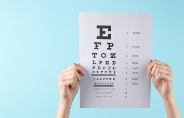 Hands holding eye test chart paper sheet on blue background. Vision correction, visit to...