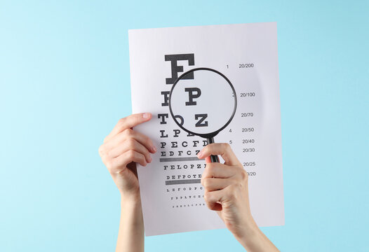 Hands Holding Eye Test Chart Paper Sheet And Magnifying Glass On Blue Background. Vision Correction, Visit To Ophthalmologist