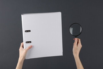 Female hand holding white binder folder and a magnifying glass on a dark background. Archive,...