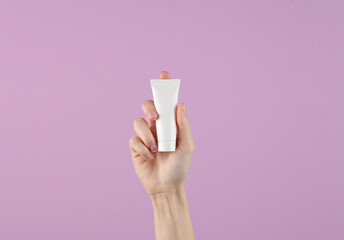 Female hand holding white cream tube on purple background. Template for design, creative beauty mockup. Skin care