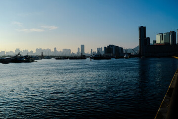 Naklejka premium The calm seascape with skyscrapers, urban skyline in the background under the sunset. Hong Kong city view with boats on the sea. Travel scene, city scene and cityscape.