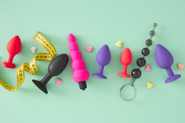 Concept of size options in adult sex toy selection. Top view arrangement different anal butt plugs and anal beads measuring tape and colorful hearts on turquoise background. - obrazy, fototapety, plakaty