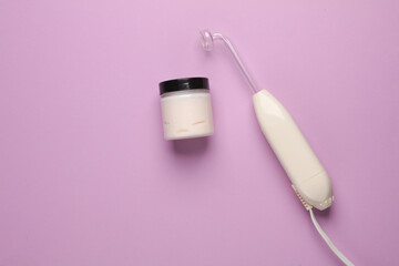 Acne treatment. Device for local darsonvalization with cream jar on a purple background. Beauty concept. Flat lay.