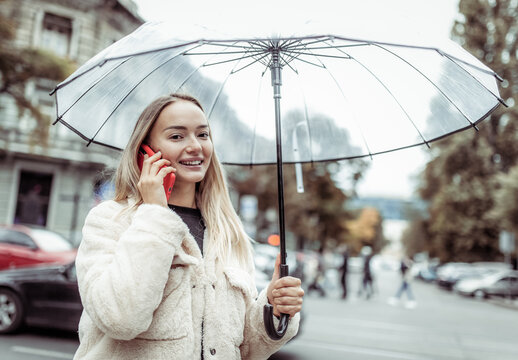 Smiling fashion blonde woman with transparent umbrella talking on phone in the city. Lifestyle, rainy weather