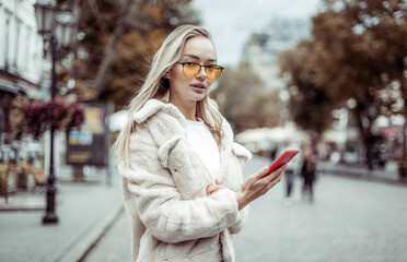 Stylish fashion woman in a fashionable fur coat and yellow glasses uses a smartphone in the city