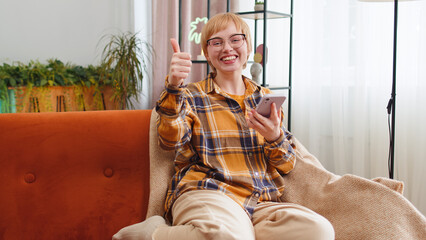 Happy young woman use mobile smartphone typing browsing say Wow yes found out great big win good news celebrate lottery jackpot doing winner gesture. Portrait of girl at home in room sitting on sofa
