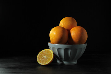 still life , closeup of a bowl with oranges and one orange cut in half on its side, on wooden table and black background