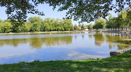 Lake in city public square. Beautiful water landscape. Park by Becket Pond in Moscow. Sunny summer day. Trees and sky are reflected in water of reservoir. Nature for recreation in middle of metropolis