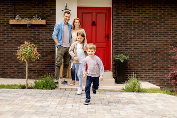 Mother and father standing outside on the front porch of a brick house, while son and daughter...
