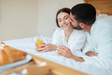 Obraz na płótnie Canvas Husband Kissing Wife As Couple Enjoying Orange Juice In Hotel