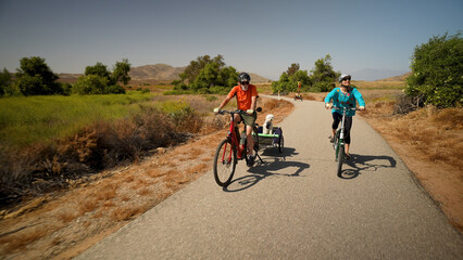 Elderly senior man biking on an electric bike on a trail pulling a trailer with a dog in it with a mature woman on an e-bike next to him.