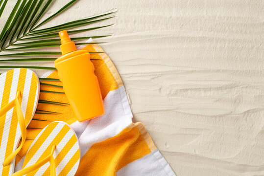 Sun protection on vacation concept. Top view photo of beach towel, yellow flip-flops, sunscreen spray and palm leaf on the sand on isolated background with copyspace