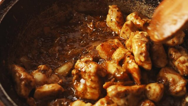 Chicken pieces in spices fried in a cast-iron skillet are mixed with a wooden spatula. Close-up