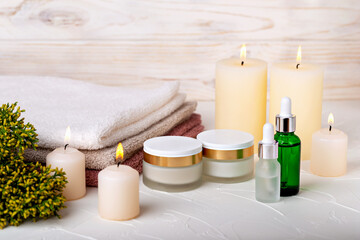 Fototapeta na wymiar Cosmetics, burning candles, towels and juniper branch on light background. Cream or lotion, oil or gel, skin care, spa treatments. Selective focus