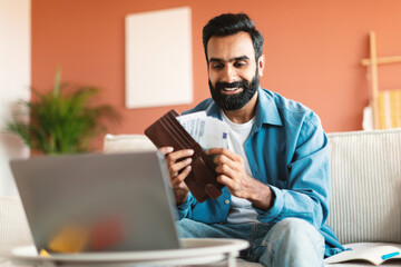 Indian Man Holding Wallet Counting Cash Near Laptop At Home