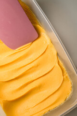 Container with mango or apricot sweet ganache on pink silicone spatula for covering and decorating cakes, macarons