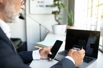 Serious european senior businessman in suit, glasses with laptop with empty screen, use smartphone, credit card