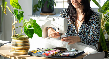 A woman sews tulle on an electric sewing machine in a white modern interior of a house with large...
