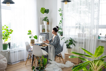 A woman sews tulle on an electric sewing machine in a white modern interior of a house with large windows, house plants. Comfort in the house, a housewife's hobby
