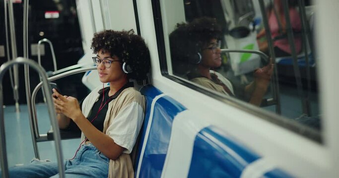 A young beautiful woman, a black African-American woman rides the subway, chats in a smartphone and listens to music in headphones. A black student rides the subway and communicates on a smartphone.
