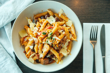 Penne pasta with olives, vegetables and parmesan cheese on a wooden table with cutlery and napkin