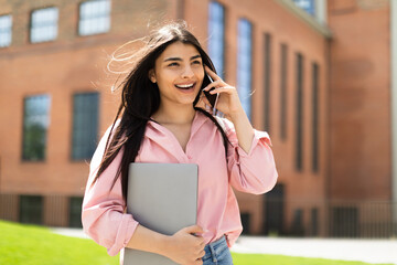 Fototapeta premium Excited hispanic lady making phone call, talking on cellphone while walking in college campus after classes, free space