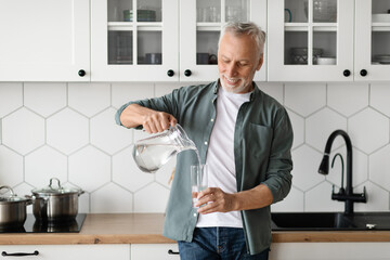 Thirsty Senior Man Pouring Water From Jug To Glass In Kitchen Interior
