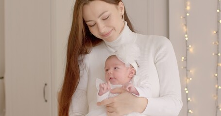 Joyful meeting of mother and child in white clothes. She smiles, holds the baby in her arms. Light lamps on the background in bokeh.