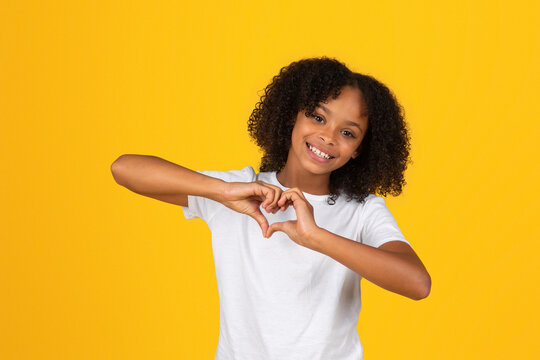 Positive Curly Teen Black Schoolgirl In White T-shirt Has Fun, Making Heart Gesture With Hands